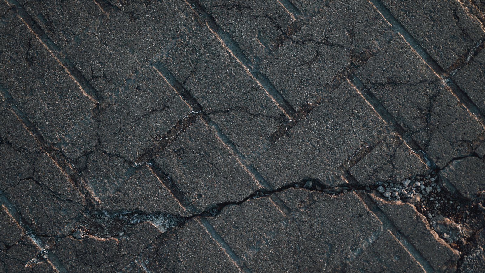 A close overhead crop of a worn grey road surface laid in diagonal rectangular slabs, split by a long jagged crack with loose gravel gathered along it, with no people, vehicles, signs or text in view.