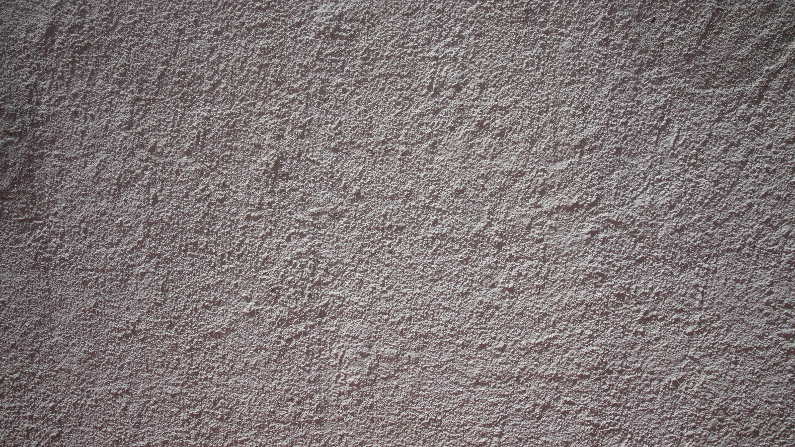 A flat, full-frame wall of coarse warm grey stucco, its raised speckled render catching light unevenly from the upper left.