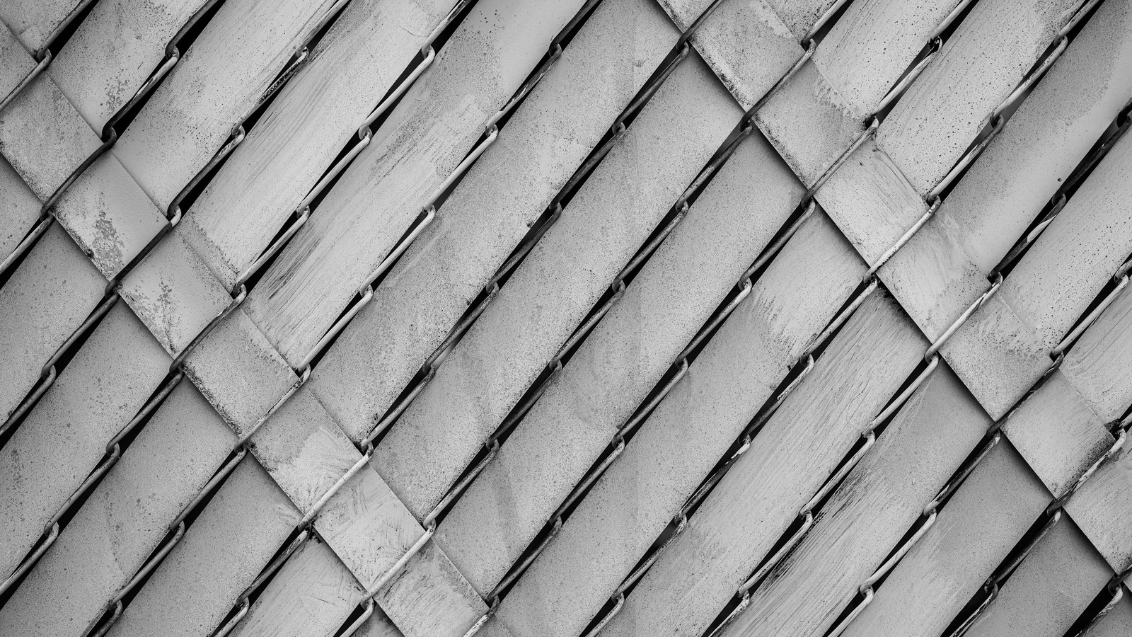 A close photograph of a chain link fence threaded with flat privacy slats, shot square on and rendered in greyscale, so the whole frame is a regular diagonal lattice of pale slats crossed by darker wire. The metal is weathered and streaked in places. No people, no lettering, no signage and no place can be identified from it.