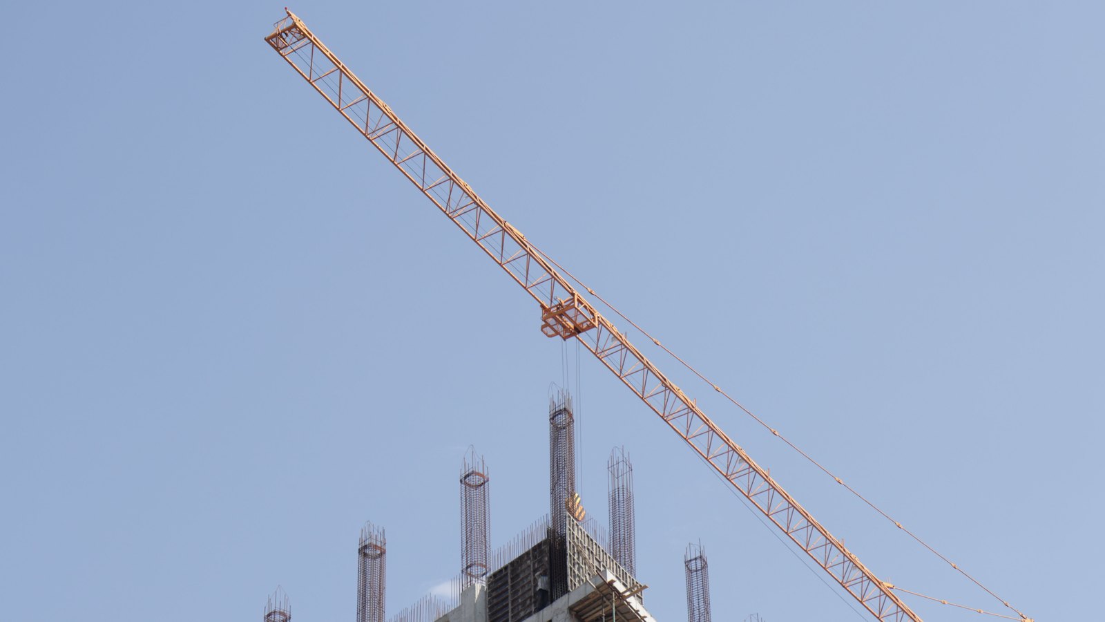 An orange tower crane rises above a concrete building under construction against a clear blue sky.