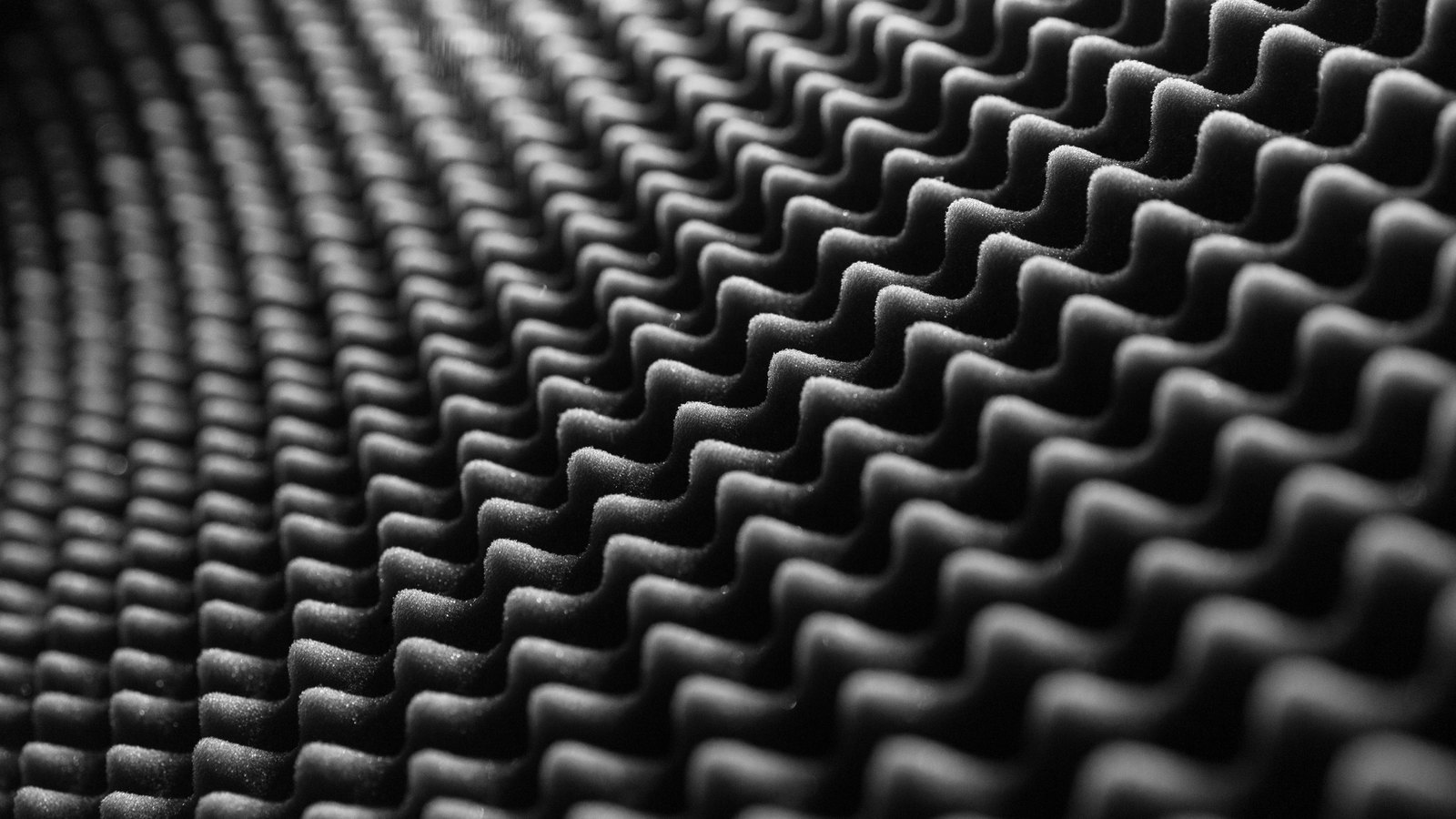 A close monochrome photograph of acoustic absorber foam, its regular egg-crate ridges running away from the camera in dark grey rows that catch a raking light along their crests and fall into shadow between.