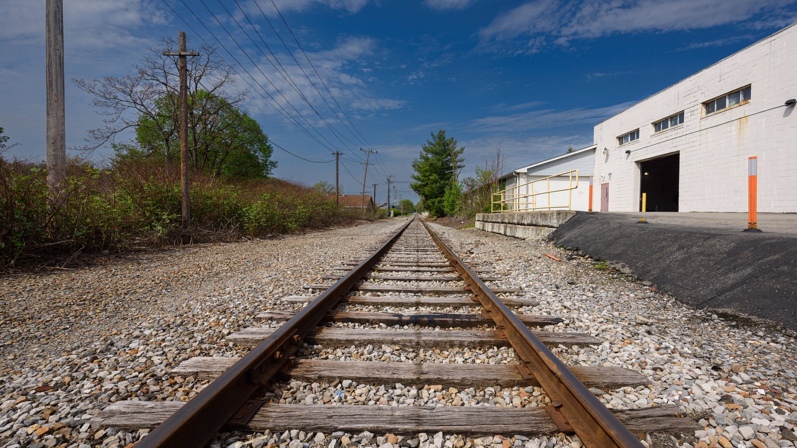 A single freight track of steel rails on weathered wooden sleepers and grey gravel ballast running away from the camera past a white blockwork warehouse with an open loading door and orange bollards, timber utility poles and overhead wires alongside, under a blue sky.