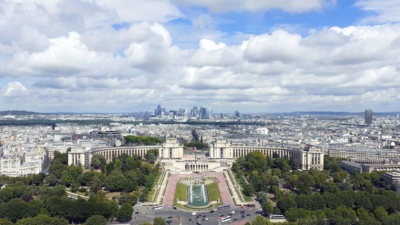 Paris seen from high above under a broad cloudy sky, the curved colonnaded wings of the Palais de Chaillot and its fountains in the foreground and the towers of the La Defense business district on the horizon. No legible text or identifiable person is in view.