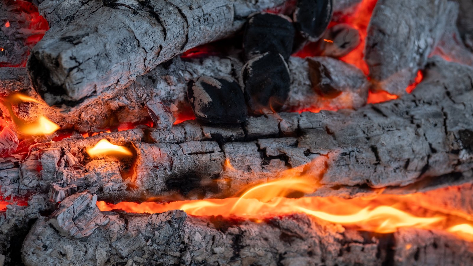 Close macro of charred logs breaking into white ash, with orange flame and glowing embers in the cracks between them.