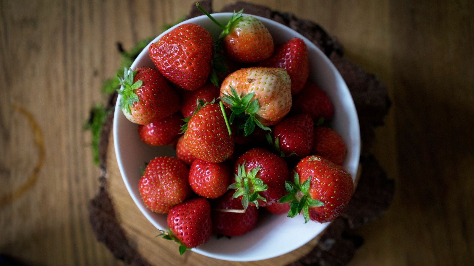 About twenty ripe strawberries piled in a plain white bowl standing on a dark wooden table, photographed from directly above. Most are deep red, a few are still pale at the shoulder, and green calyxes and stalks show between them. No packaging, label or lettering is in the frame.