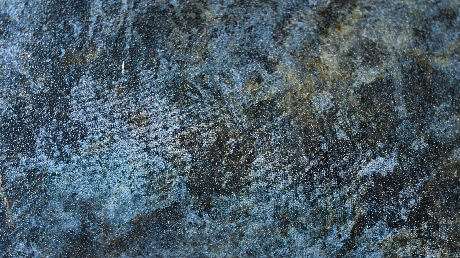 A macro view of a rough stone face in blue grey, its mottled surface flecked with pale mineral grain and darker olive patches.