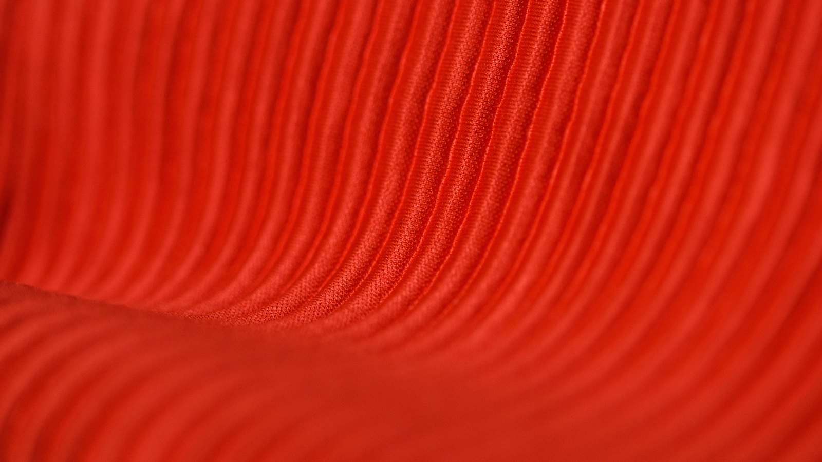 A close photograph of red ribbed corduroy, the cord lines running down from the top of the frame and curving away to the left across a soft fold, the weave visible along the raised ribs and the lower half falling out of focus. No text, lettering, brand mark, object, person or place is in view.