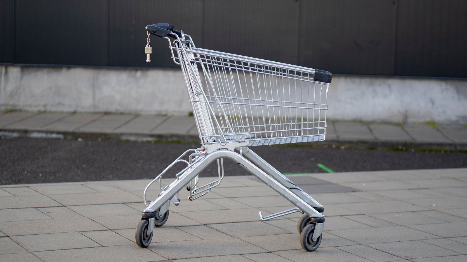 A single empty wire shopping trolley standing alone on paving slabs in front of a plain dark wall, its basket empty and its wheels turned. No brand mark, logo or readable word is in the frame and there are no people.