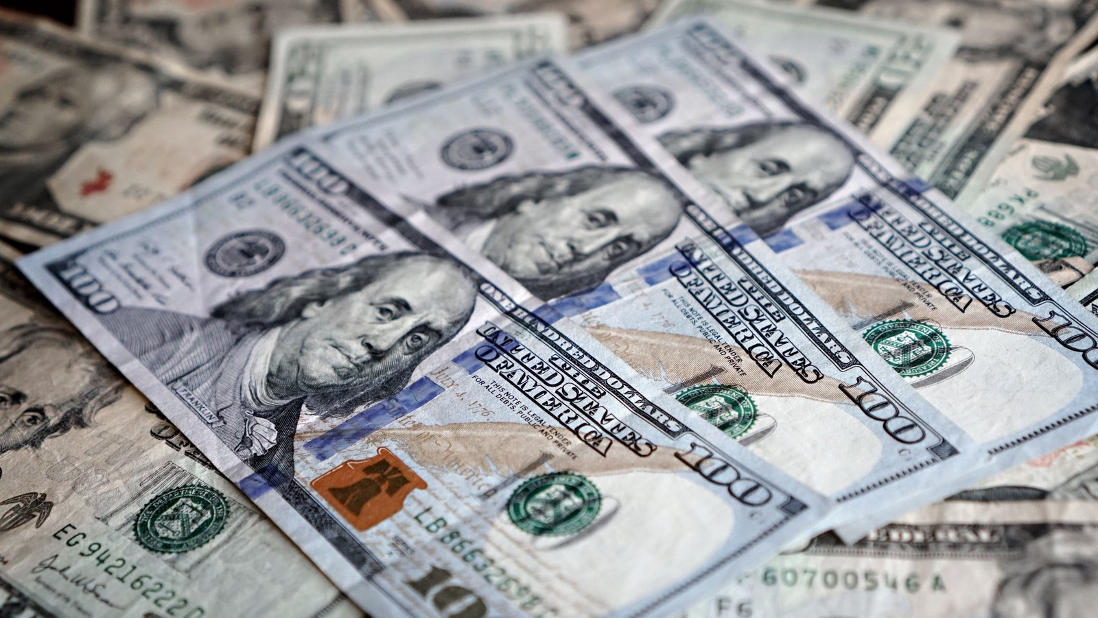 Overlapping United States one hundred dollar bills spread across the frame, with twenty dollar notes beneath them.