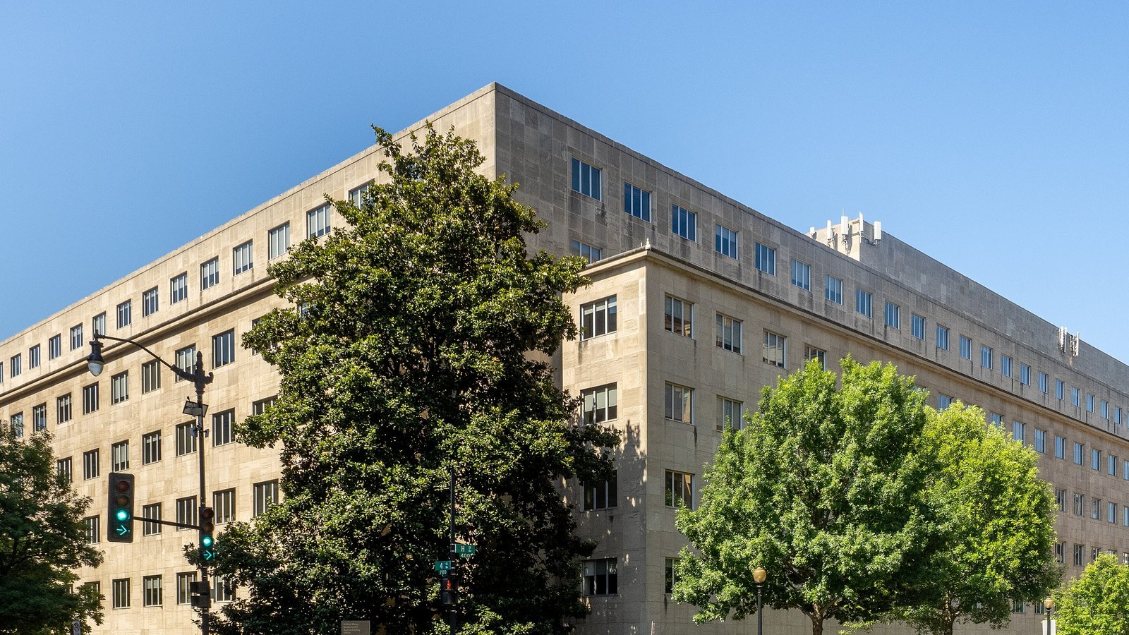 The United States Government Accountability Office building in Washington photographed from the street corner on a clear day. A five storey limestone block runs away to the left and to the right of the corner, with regular rows of small square windows across both elevations and a plain parapet against a deep blue sky. Mature green trees stand along both pavements, a green street blade on a pole reads H Street North West, and a low granite sign at the foot of the corner carries the agency's name.