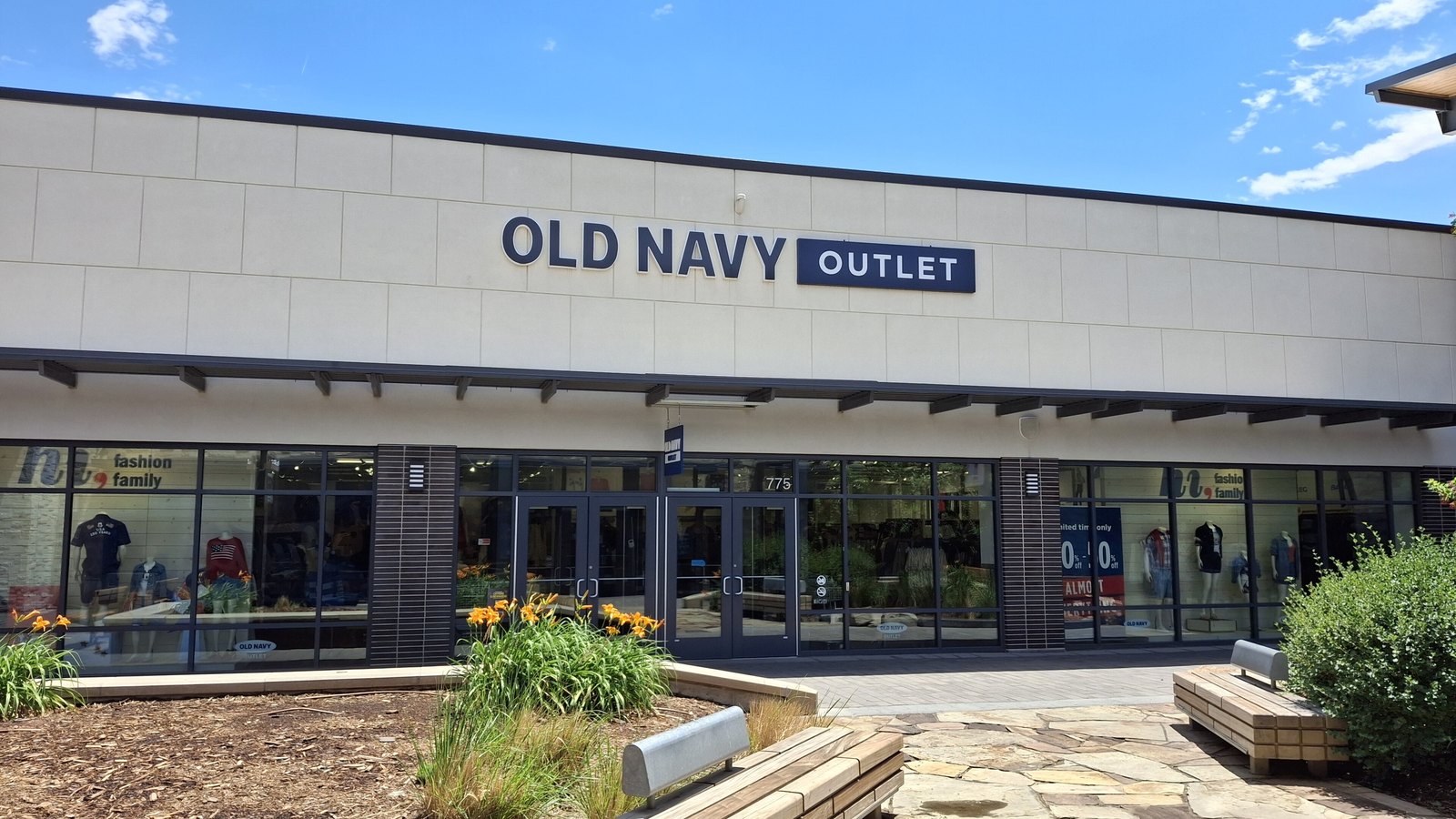 An Old Navy Outlet store at Denver Premium Outlets in Colorado, photographed in June 2026 under a bright blue sky: a low cream stone building with OLD NAVY OUTLET in navy capitals across the fascia, a glazed shopfront with dressed mannequins and a discount banner behind the glass, and day lilies in a planting bed in front. No shoppers are present.