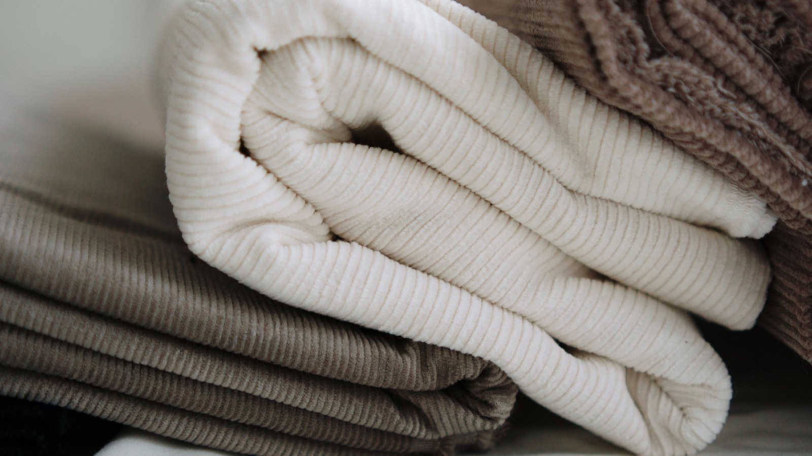 A close view of folded lengths of corduroy in cream, taupe and dark brown, stacked one on another so that the fine parallel ribs of the cloth run across the whole frame. No label, text, brand, person or place is visible.