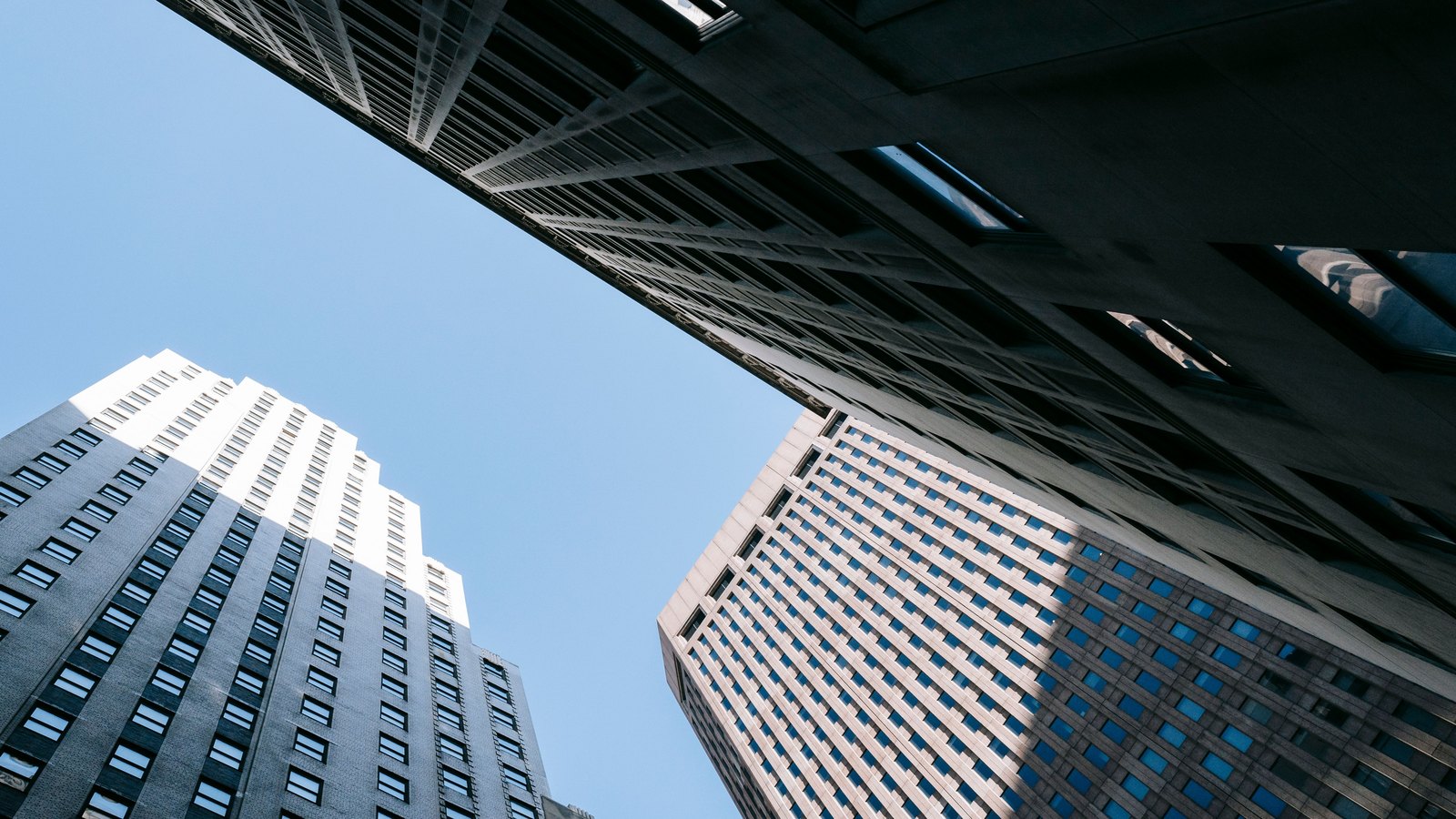 Three office towers photographed from street level looking straight up, their facades converging against a clear blue sky. One is dark stone with rows of small square windows, one is pale masonry with a grid of blue-tinted glass, and a third overhangs the top of the frame. No lettering, sign or logo is visible anywhere in the picture.