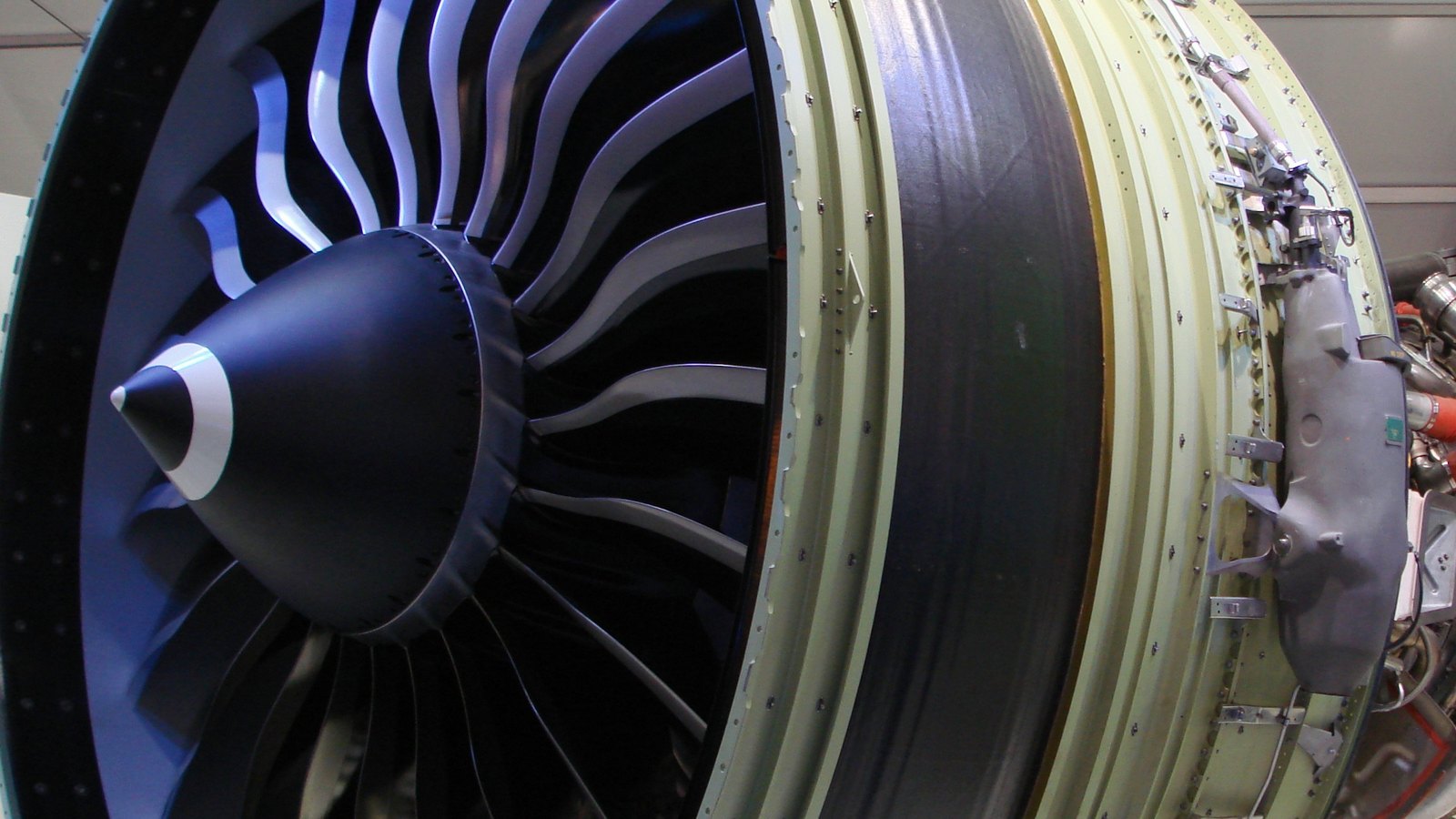 Close view of a General Electric GE90 turbofan on a display stand, showing the dark spinner, curved composite fan blades and the pale green fan case with fittings along its side.