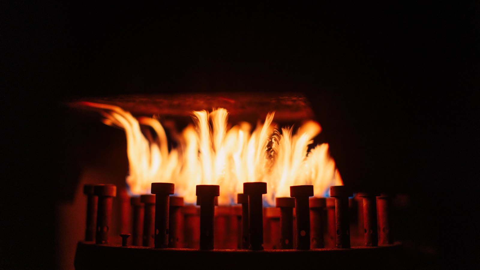 Rows of gas burner jets alight inside a dark housing, orange flames rising against a heat plate above.