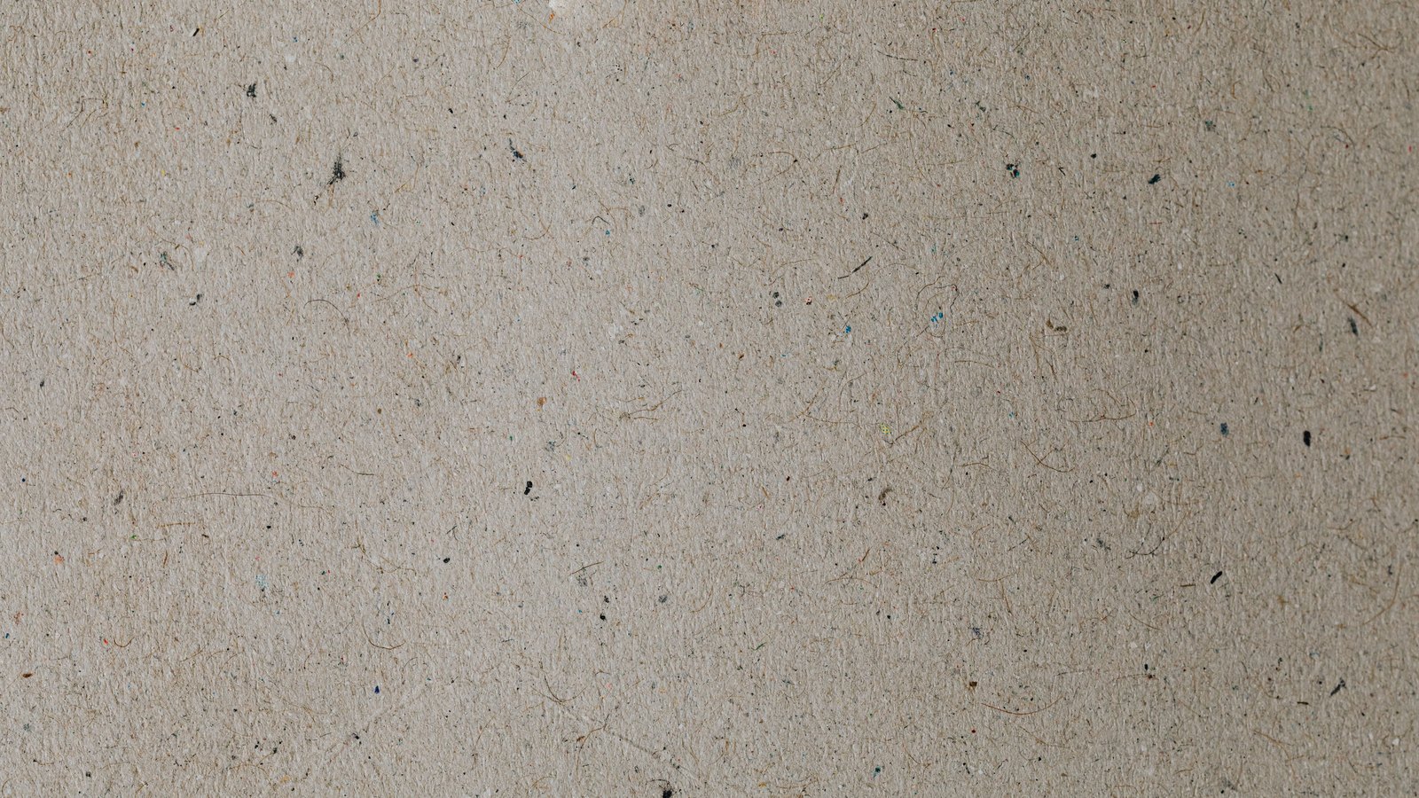A flat sheet of warm grey recycled paperboard filling the whole frame, its fibrous surface flecked with short brown fibres and scattered specks of black, blue and orange.