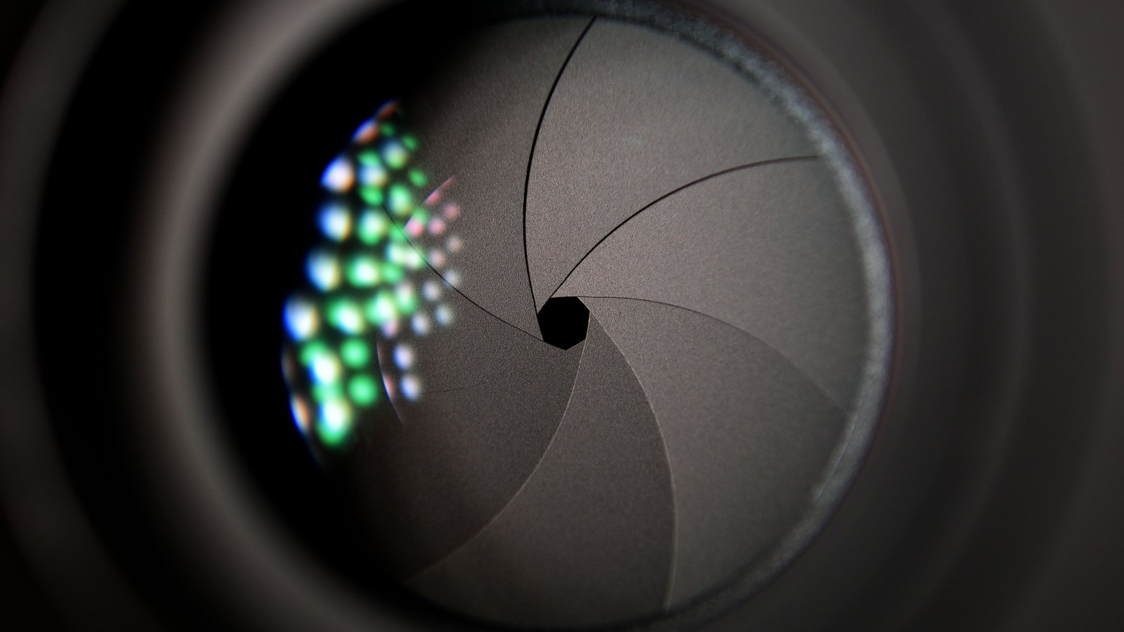 Close-up of a camera lens with its dark grey aperture blades closed down to a small opening, coloured reflections on the glass at left.