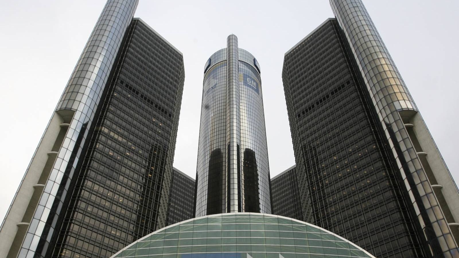 The glass towers of the Renaissance Center in Detroit seen from below against a pale overcast sky, a GM logo on the central tower and the curved glass roof of the entrance atrium in the foreground.