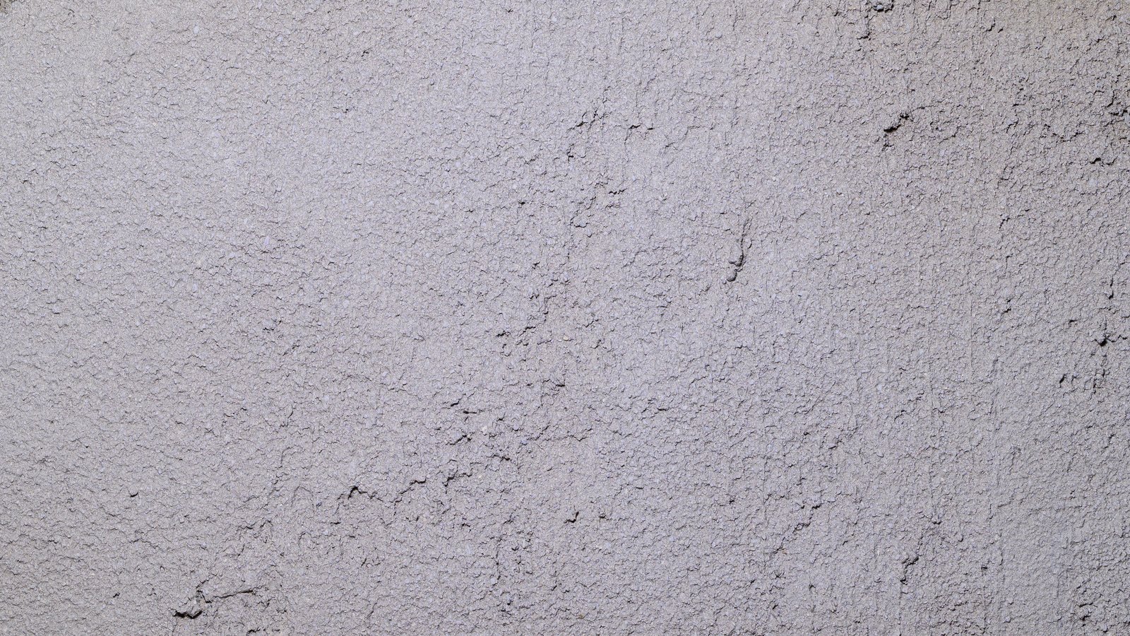 A close view of a pale grey rendered wall filling the whole frame, its coarse granular surface crossed by a few fine dark hairline cracks and shading slightly darker towards the right edge, with no people, buildings, signs or text in view.