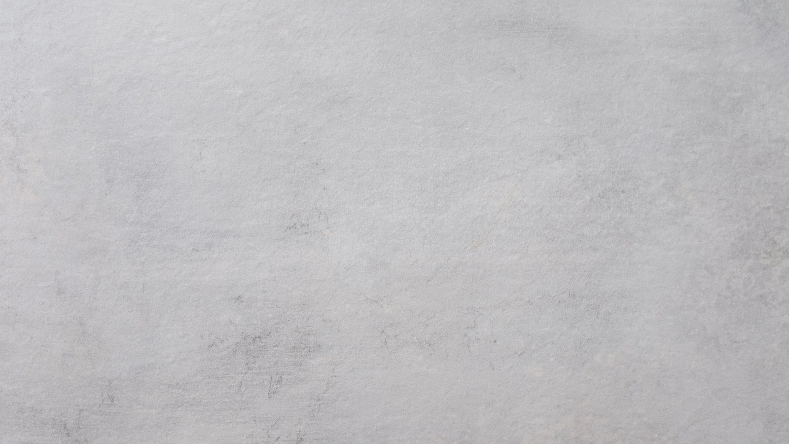 A flat expanse of pale grey concrete filling the whole frame, lit evenly and photographed straight on, its surface faintly mottled with lighter and darker patches and crossed here and there by hairline marks and a few small darker flecks near the lower edge. No people, faces, vehicles, buildings, rooms, signage or lettering are visible and no place can be identified from it. A plain surface used as neutral illustration, not a photograph of any person, place or event in this item.