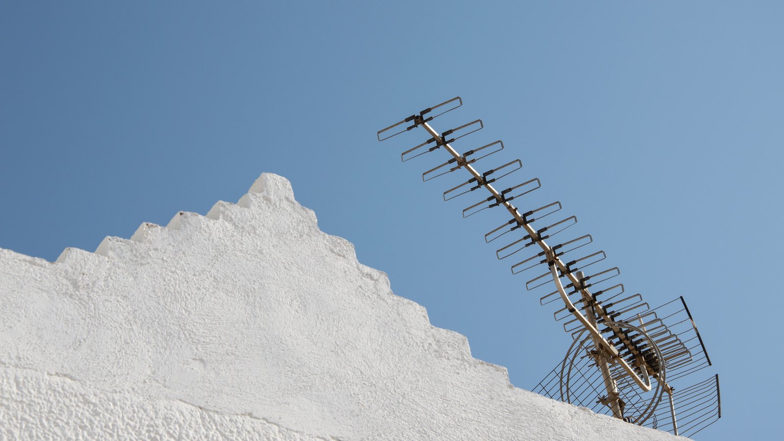 A weathered metal television aerial mounted on the stepped parapet of a whitewashed roof, its rows of short elements angled up against a clear blue sky.
