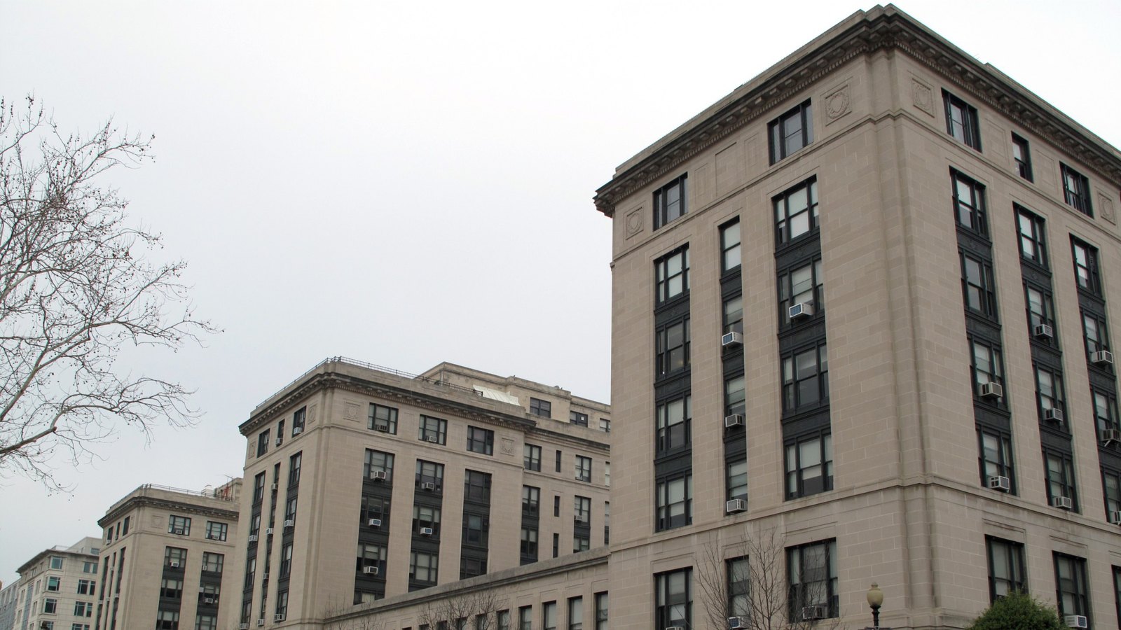 A photograph of the limestone facade of the United States General Services Administration headquarters in Washington under a flat overcast sky, a tall corner block of seven storeys with dark window frames and window air conditioning units, a lower wing receding to the left and a bare winter tree at the edge of the frame. No people, vehicles, text, signage or logo are in view.