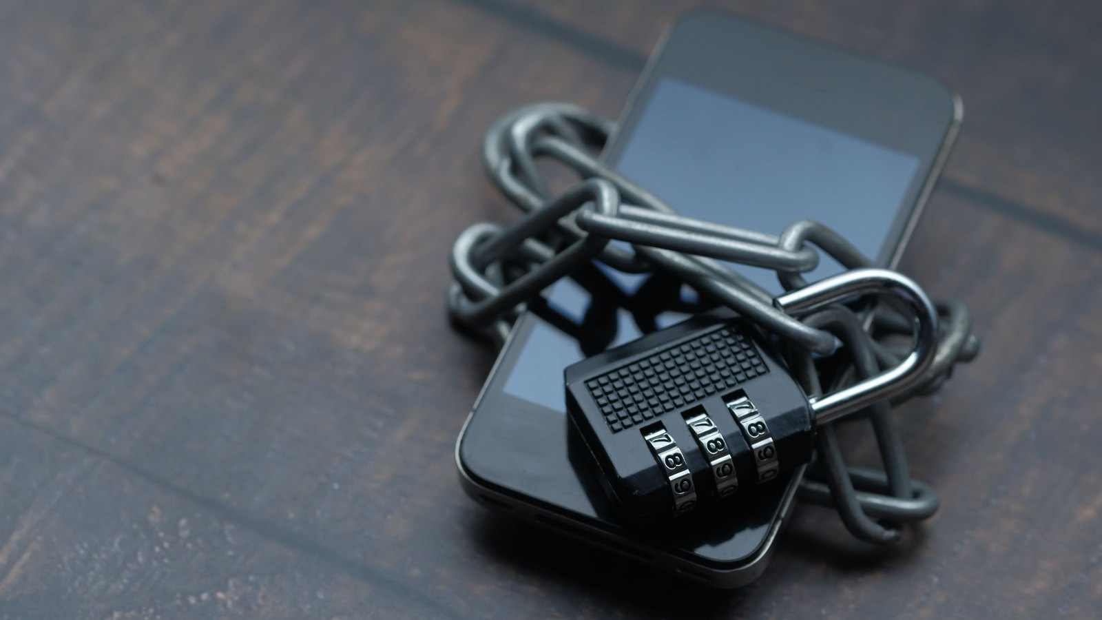 A black smartphone is wrapped in a heavy chain secured by a black combination padlock.