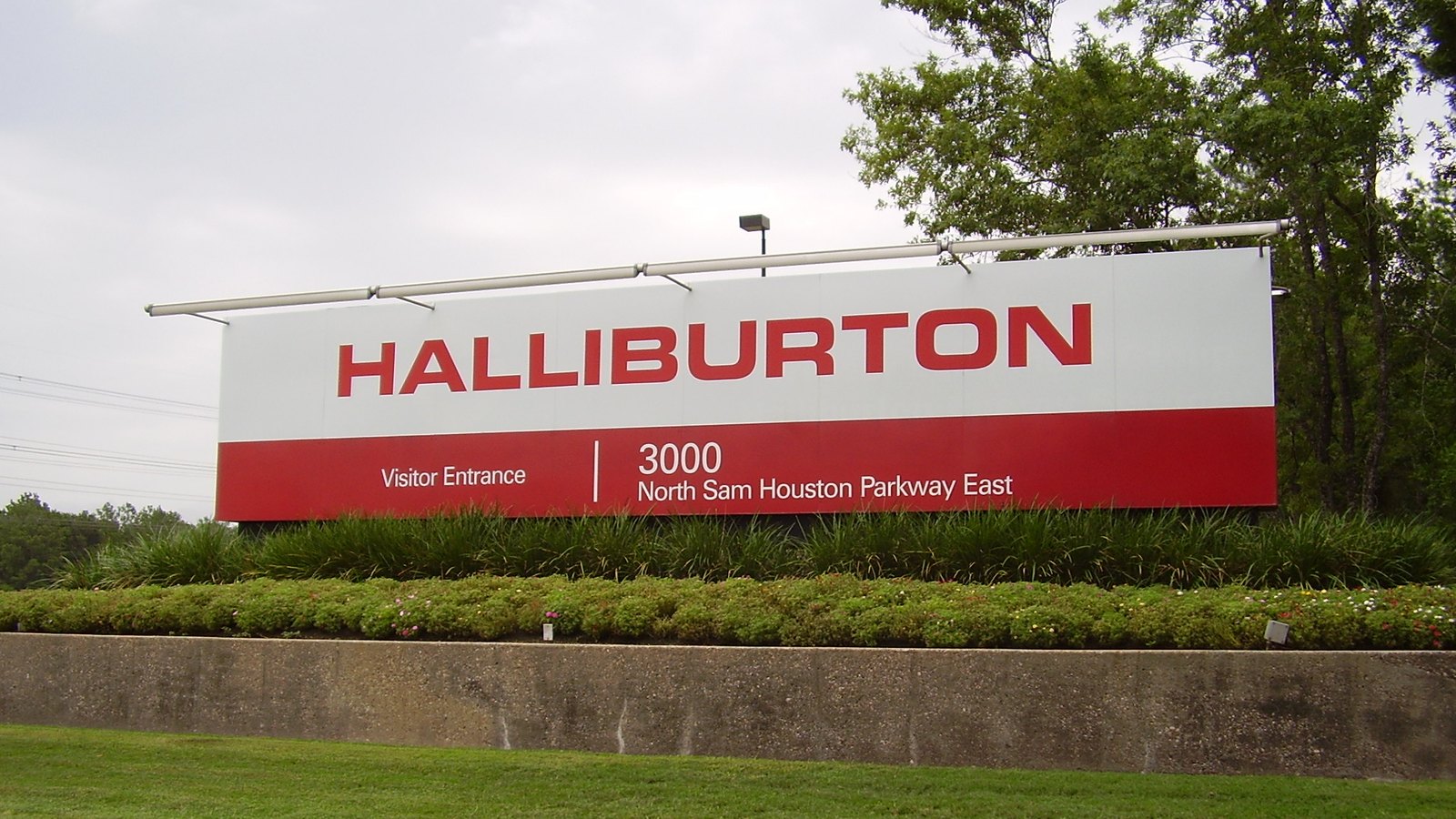 A long red and white Halliburton sign at the visitor entrance of the company's North Belt offices in Houston, set above a planted bed and a low concrete wall, with trees behind and an overcast sky.