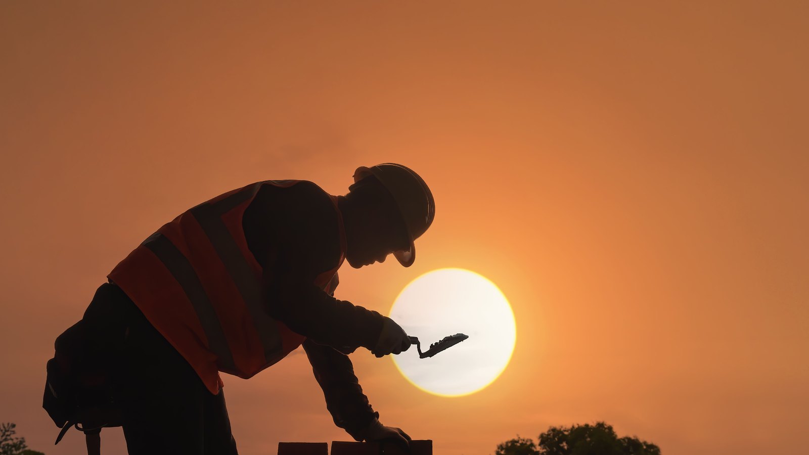 The silhouette of a construction worker bent over a course of bricks with a trowel, hard hat and high visibility vest outlined against a low white sun in an orange sky, a dark tree line along the horizon behind. No face, signage, plate, logo or landmark is identifiable. It illustrates outdoor work carried on through the hot part of the day and is not a photograph of any Lee or Collier County worksite.