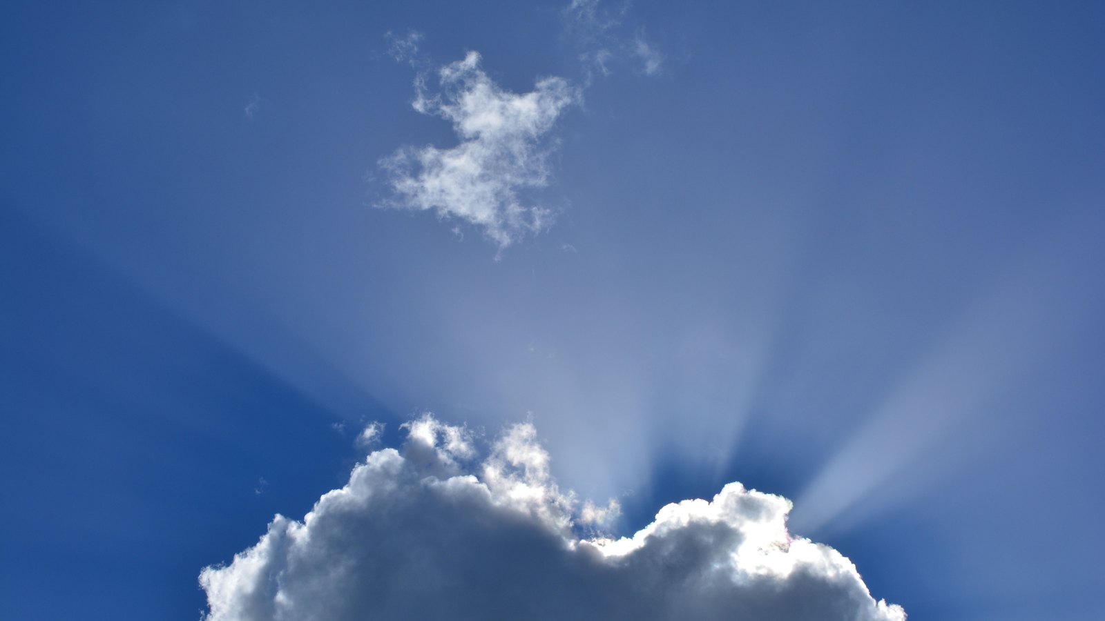 A deep blue sky filling the frame, with the top of a white cumulus cloud rising from the bottom edge and the sun hidden directly behind it, throwing a fan of pale rays upward and outward across the blue. A small wisp of thin cloud sits higher and left of centre. No ground, horizon, buildings, people, signage or text are in view.