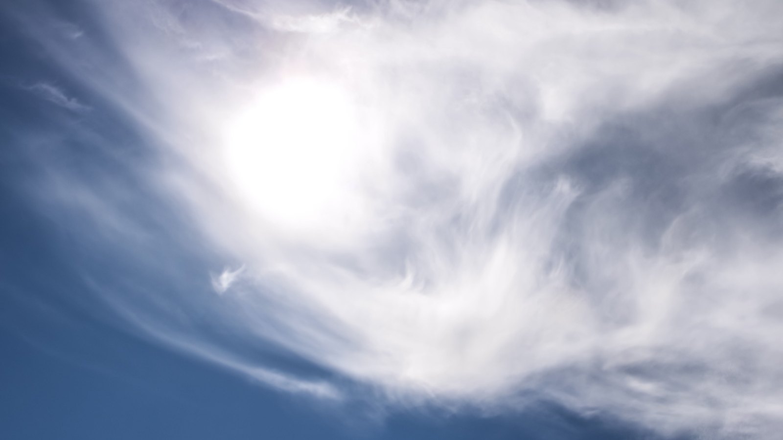 Looking straight up at the sun. It burns as a hard white disc, over-exposed at the centre, behind a thin sheet of wispy cirrus cloud that streams across the frame in feathered curls. The sky is deep blue at the lower left and washes out to pale grey around the sun. No ground, horizon, buildings, people, signage or text are in view.