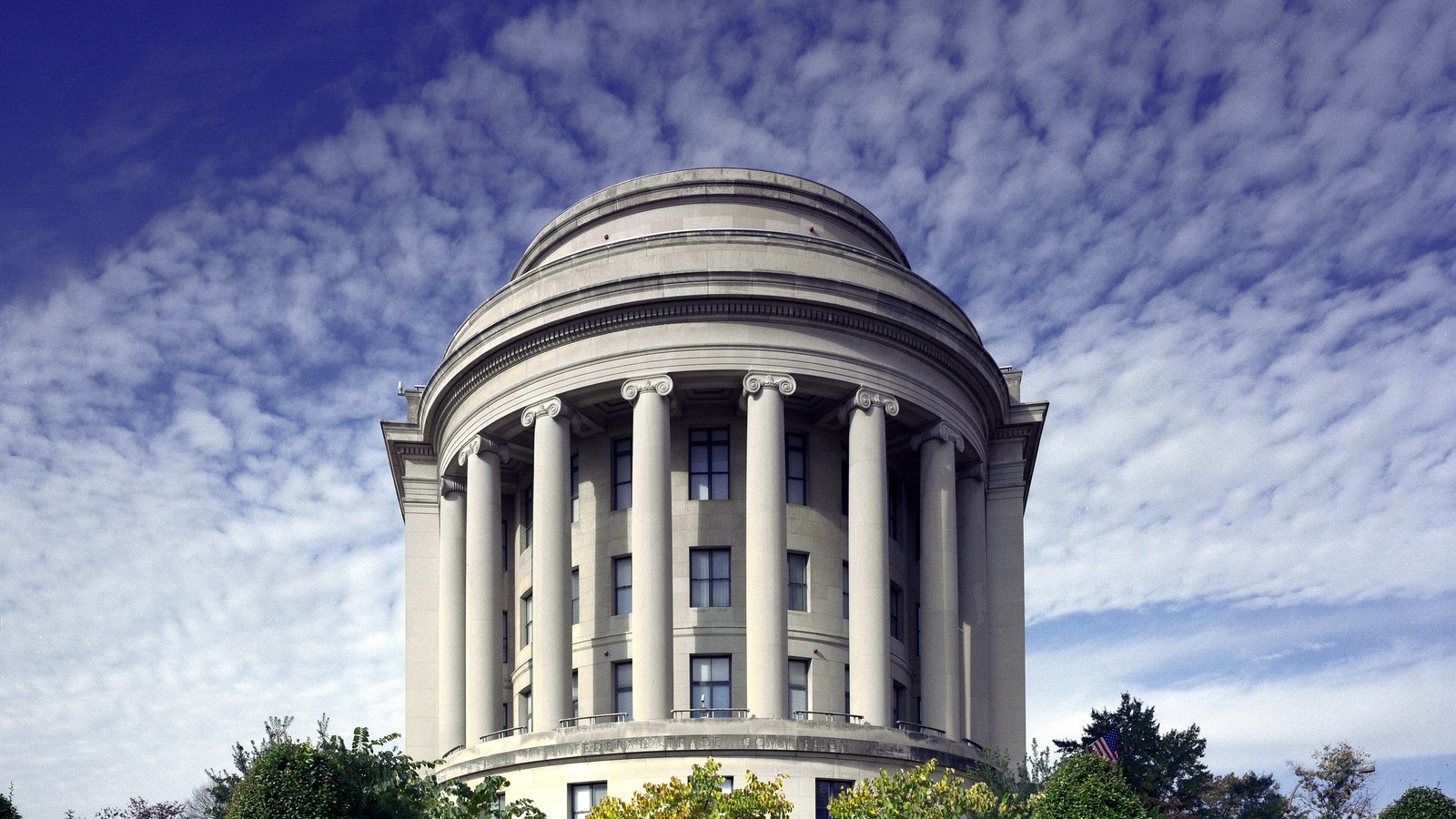 The curved colonnaded end of the Apex Building in Washington, headquarters of the Federal Trade Commission, photographed straight on. A row of tall Ionic columns runs around the rounded facade beneath a shallow drum, with clipped trees and shrubs across the foot of the building and a sky of small scattered clouds behind it.