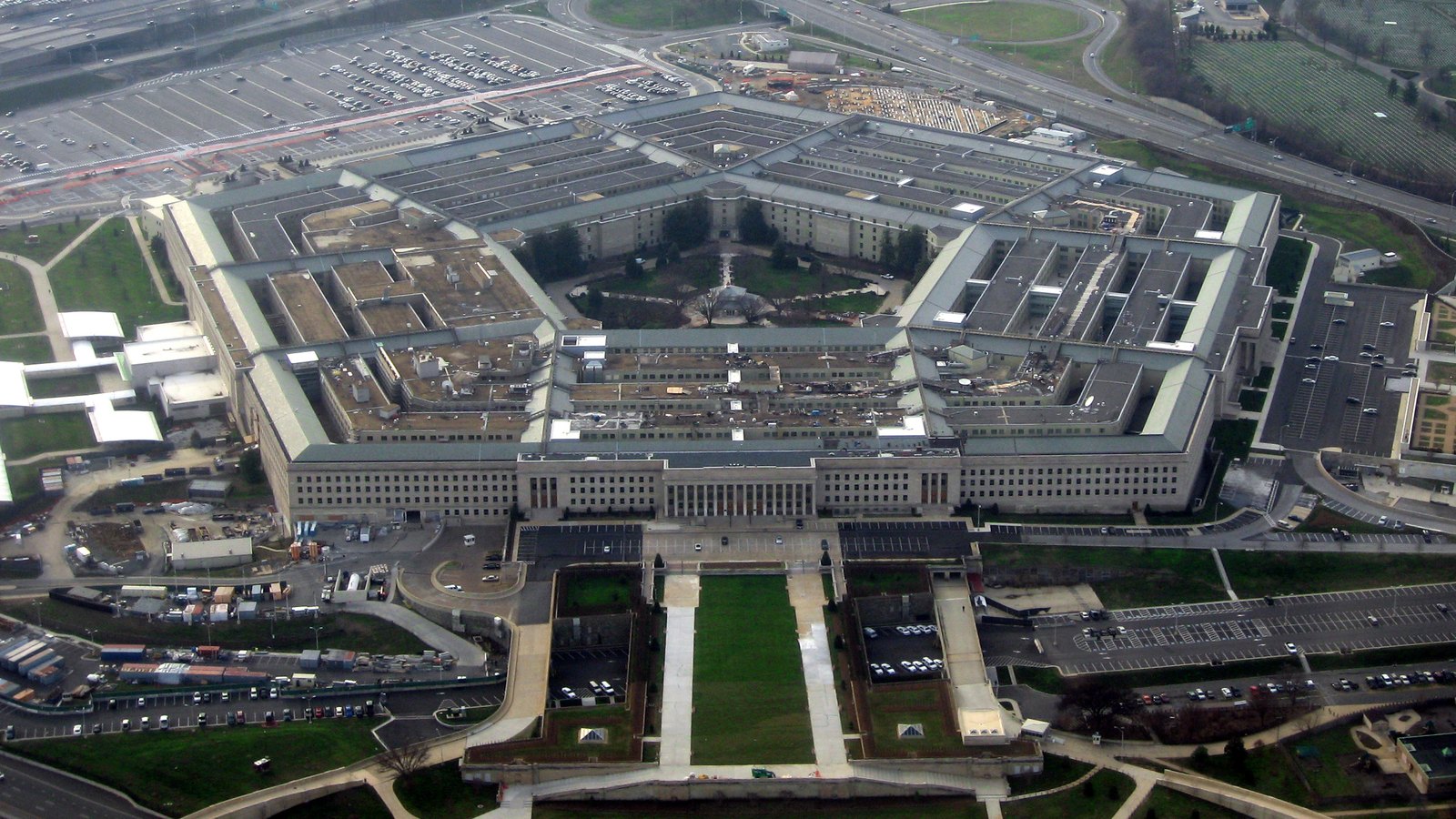 An aerial photograph of the Pentagon in Arlington, Virginia, taken from the air on a clear winter day. The five sided building fills the centre of the frame, its concentric rings and central courtyard visible, with roads, car parks, bare trees and mown lawns around it. Nothing at this distance is identifiable as a person or a particular vehicle.