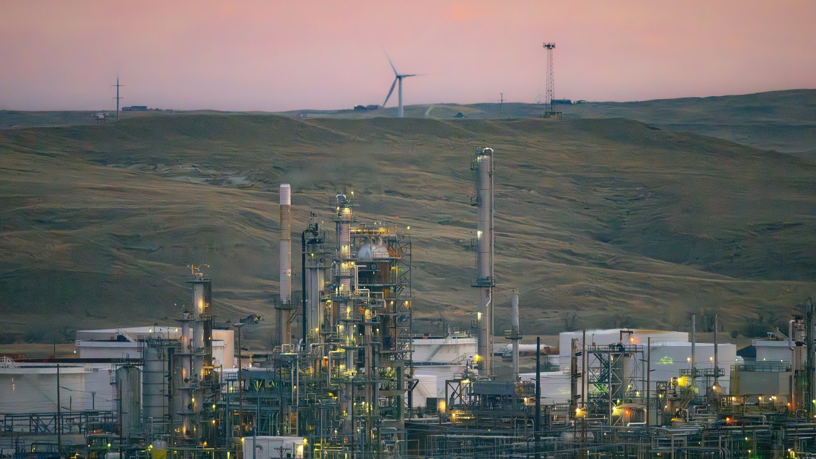 An oil refinery at dusk below a bare rolling Wyoming hillside. Distillation columns, stacks, pipework and rows of white storage tanks are picked out by yellow work lights, a single wind turbine and a lattice tower stand on the ridge above, and the sky behind is pink. No signage, lettering or people are visible.