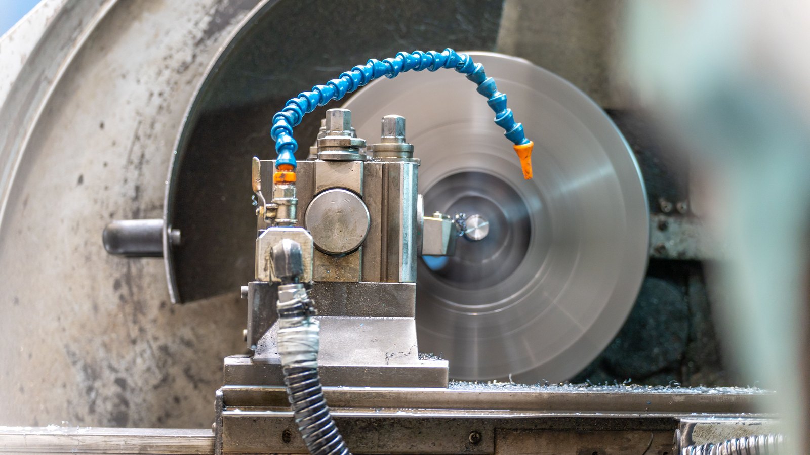 A close view of a lathe cutting a spinning steel workpiece, two blue segmented coolant hoses arched over the tool post and bright swarf collected on the machine bed.