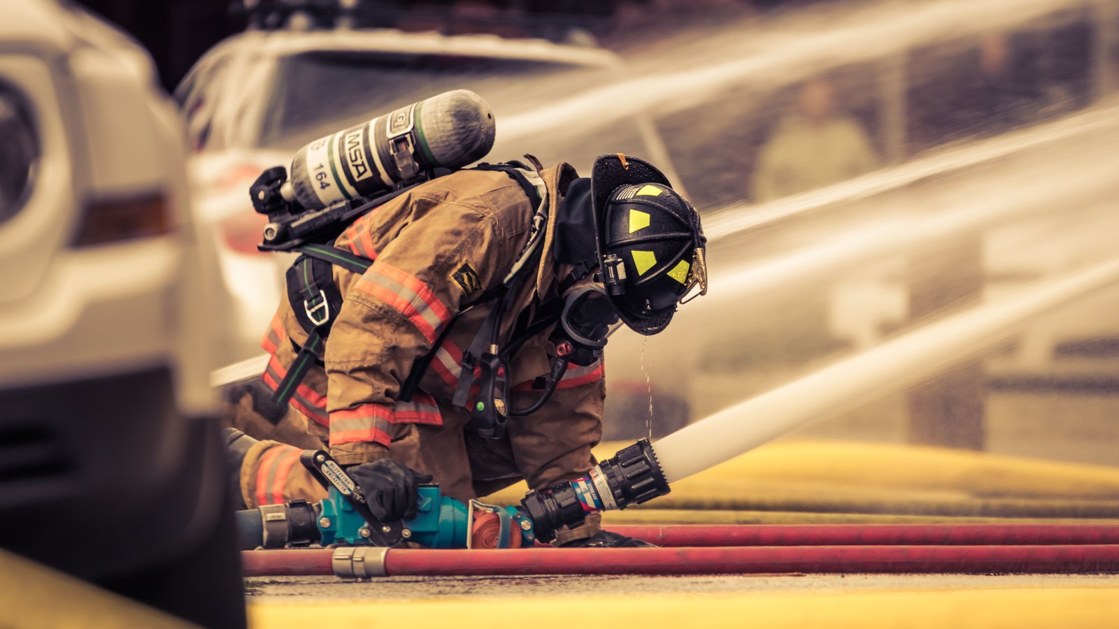 A firefighter in tan turnout gear kneels over a hose line, working a valve, with water arcing across the frame behind. The face is fully covered by a breathing mask and helmet, and the only lettering anywhere is the air cylinder maker's initials and a cylinder number. No vehicle livery, number plate, street sign, house number or department marking appears in the frame. Stock photograph, not a photograph of this event.