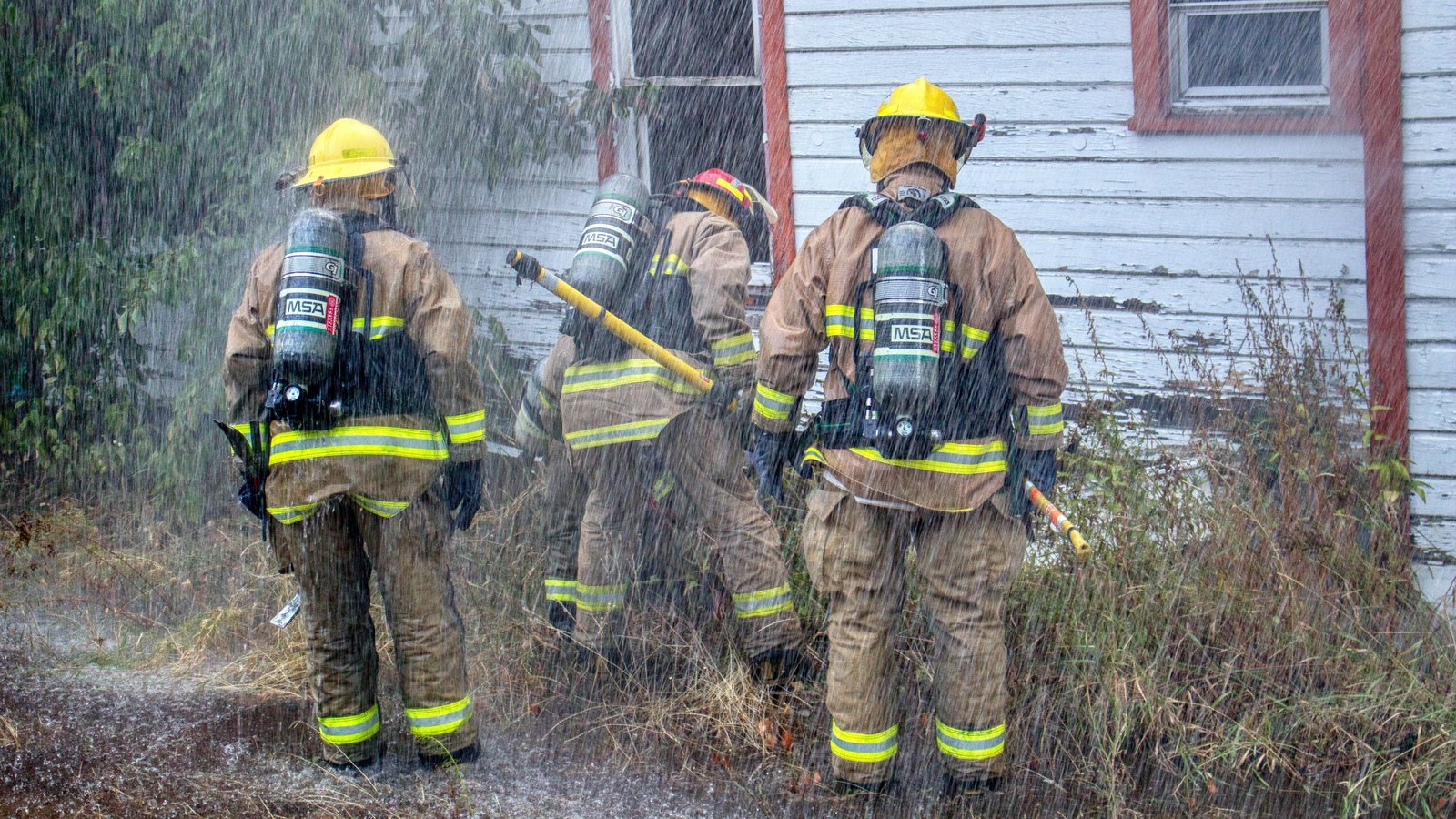 Three firefighters in tan turnout coats and yellow helmets stand with their backs to the camera beside the pale clapboard wall of a single storey house, working in a heavy curtain of falling spray. Their air cylinders are the only lettered objects in the frame and carry an equipment maker's initials rather than any department name. No faces, no vehicles, no number plates, no street signs, no house number and no agency markings appear anywhere. Stock photograph, not a photograph of this event.