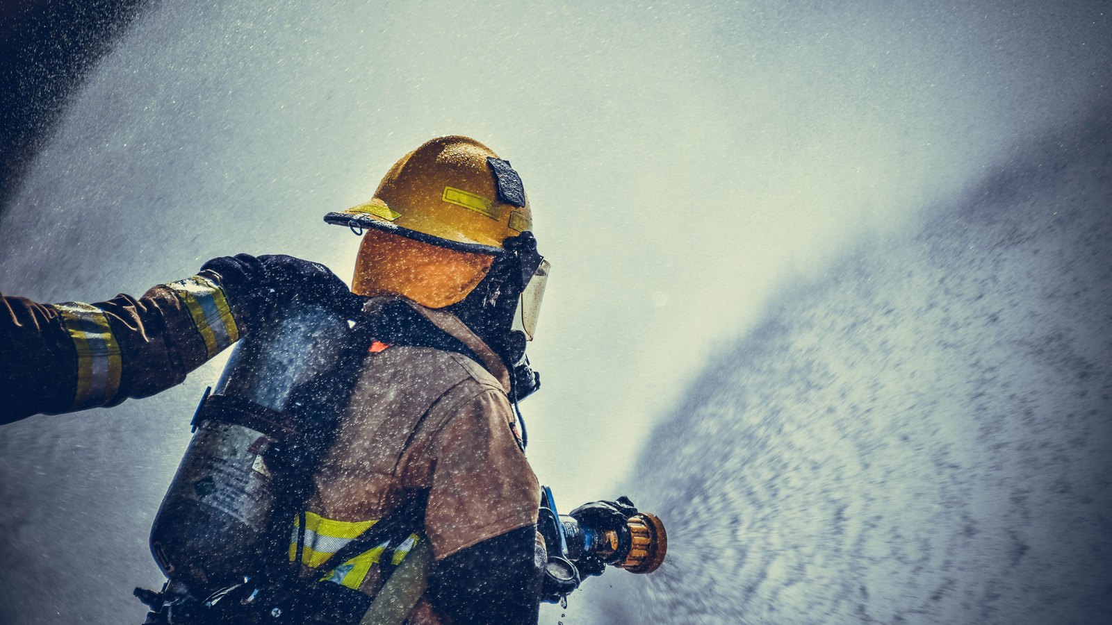 A single firefighter photographed from behind in dark turnout gear, an air pack and a yellow helmet, working into a heavy curtain of falling spray that fills most of the frame. No face is visible and there is no vehicle, number plate, building, street, signage or agency lettering anywhere in the picture. Stock photograph, not a photograph of this event.