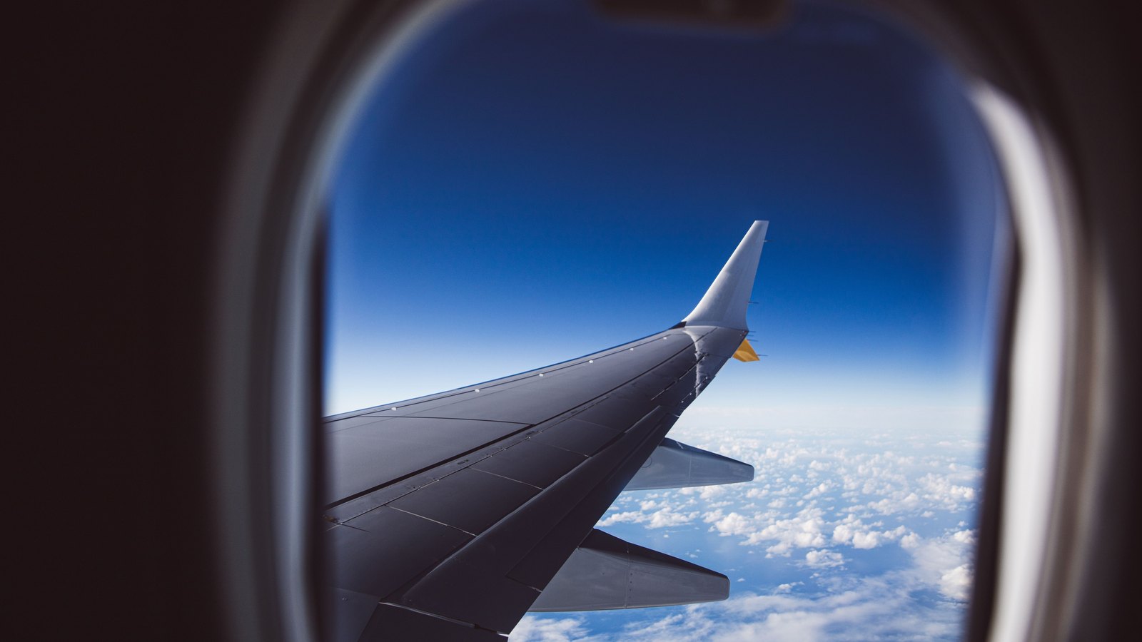 The wing of a passenger aircraft in flight, seen through a cabin window, against deep blue sky above a broken deck of white cloud. The winglet is plain and carries no airline name or marking.