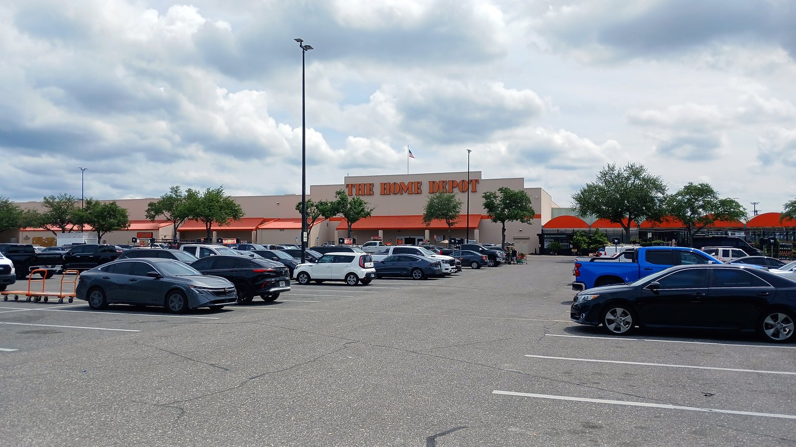 A Home Depot store seen across its car park under a cloudy sky, the company name in orange lettering along a tan wall above an orange awning, with a United States flag on a pole behind the roofline.