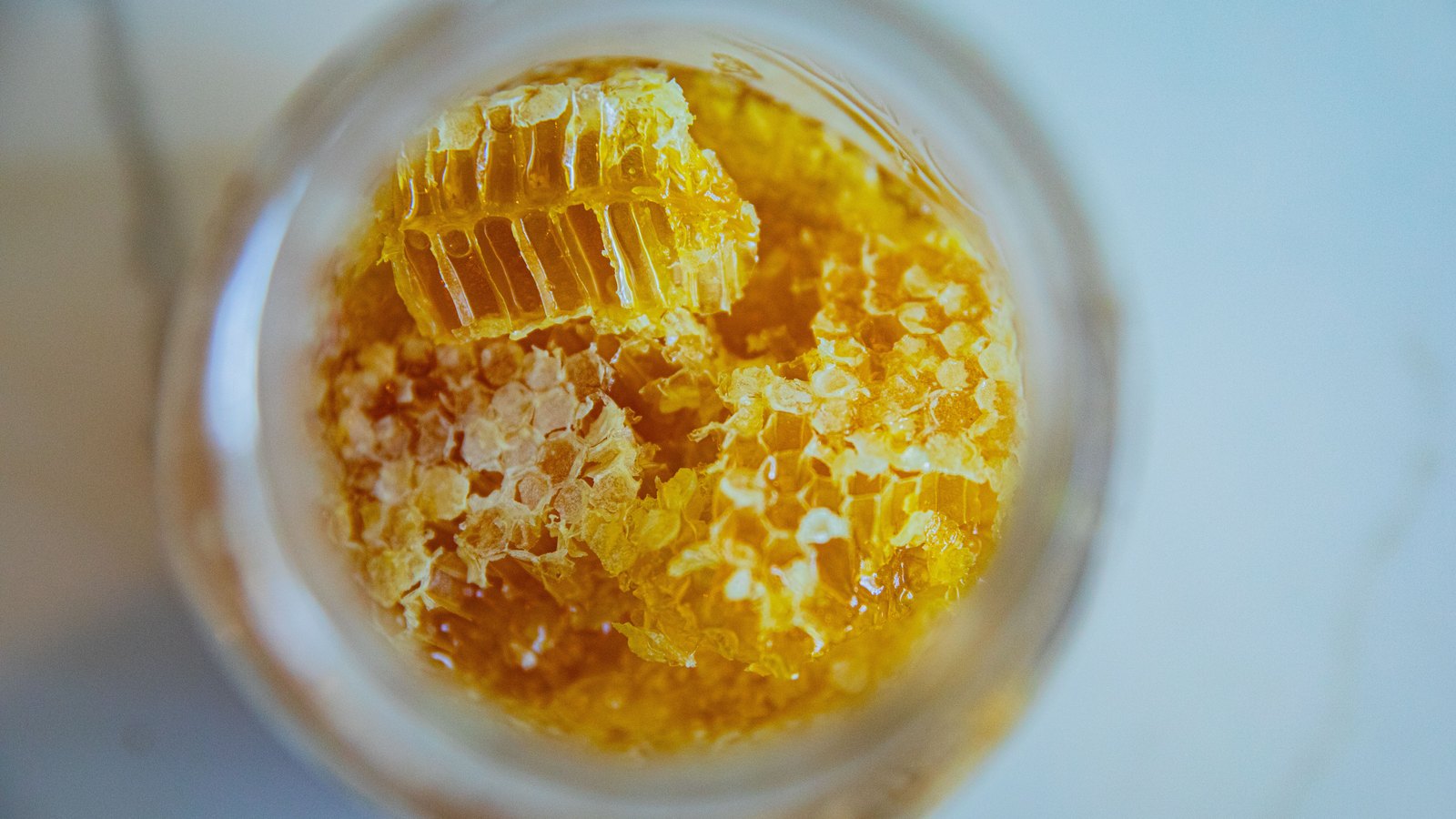 Looking straight down into the open mouth of a plain glass jar filled with honey, several irregular pieces of pale wax honeycomb floating at the surface with their hexagonal cells holding amber liquid, against a soft out of focus white background. No label, lettering or mark is on the jar.