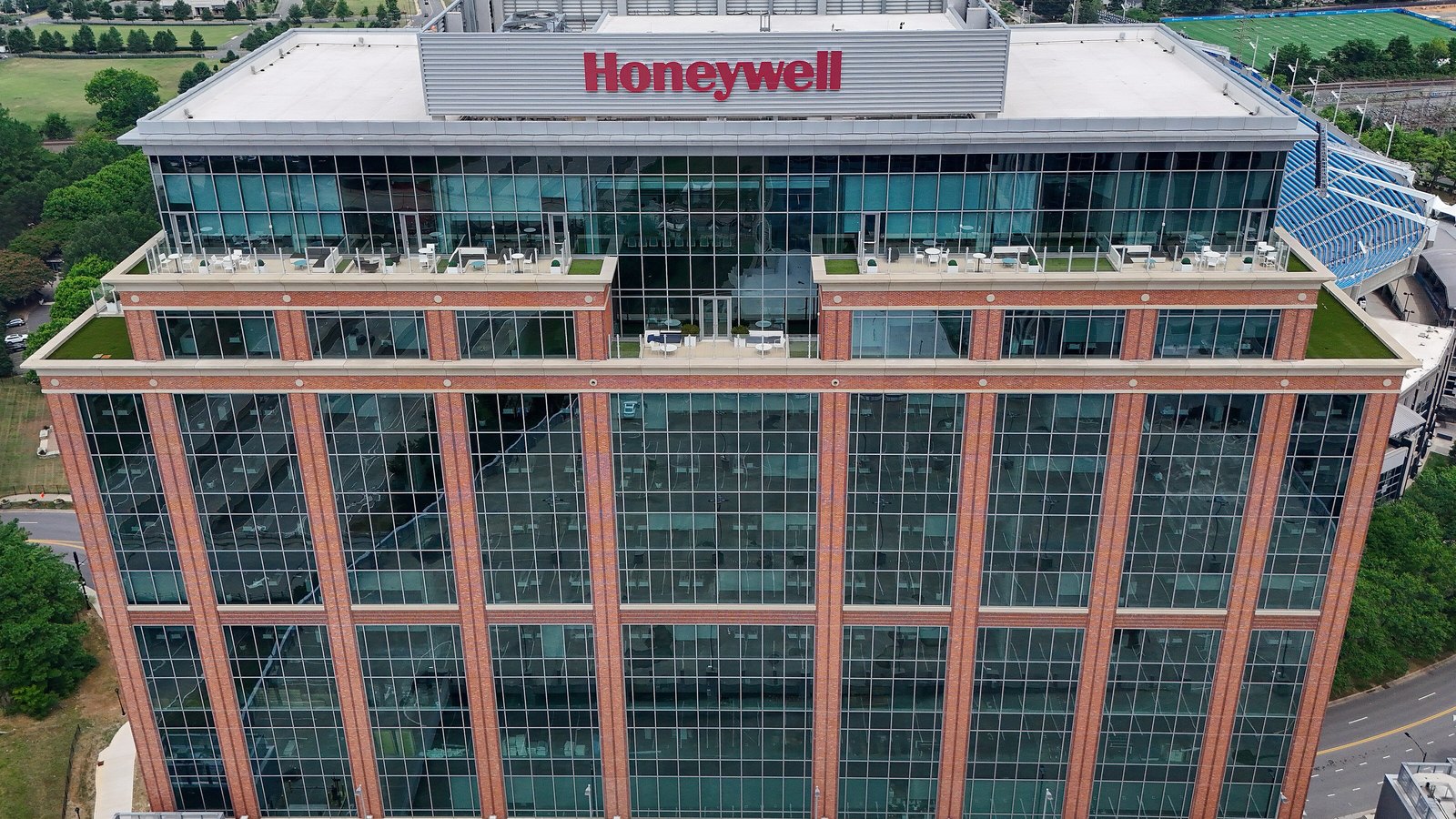 Aerial view of a brick and glass office tower with the Honeywell name in red letters on its rooftop, roof terraces with tables and chairs, and trees and a stadium beside it.