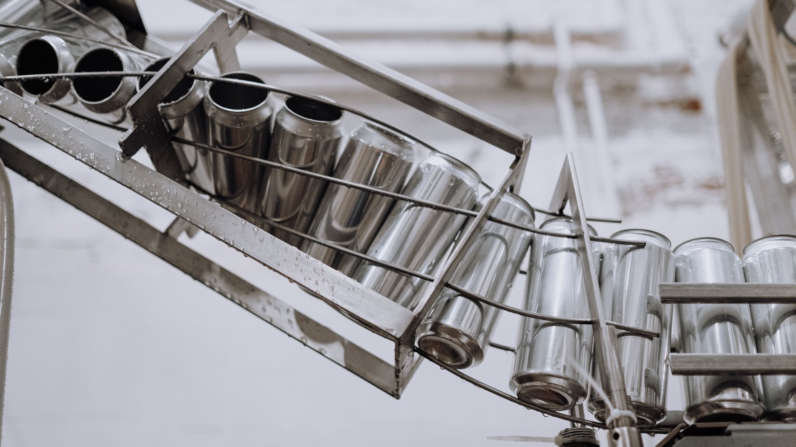 Unlabelled aluminium cans carried on their sides along an overhead stainless steel rack conveyor in a canning plant, beaded with condensation, with no people and no printing on the cans.
