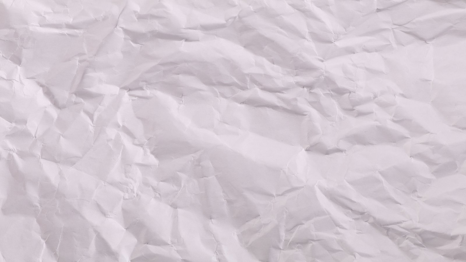 A full frame close photograph of a sheet of crumpled white paper, its creases and soft ridges picking up a light from the upper left, carrying no writing or printing.