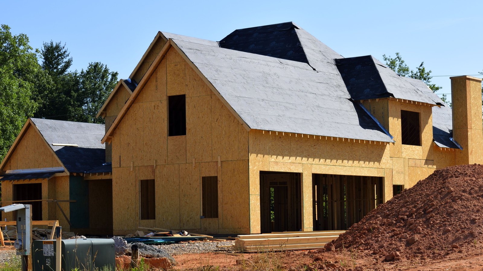 A large two storey house under construction, sheathed in oriented strand board, with dark underlayment laid over the roof and the window and door openings still open. A stack of fresh lumber and a heap of red clay soil lie in front of it under a clear blue sky. No people are in the frame.