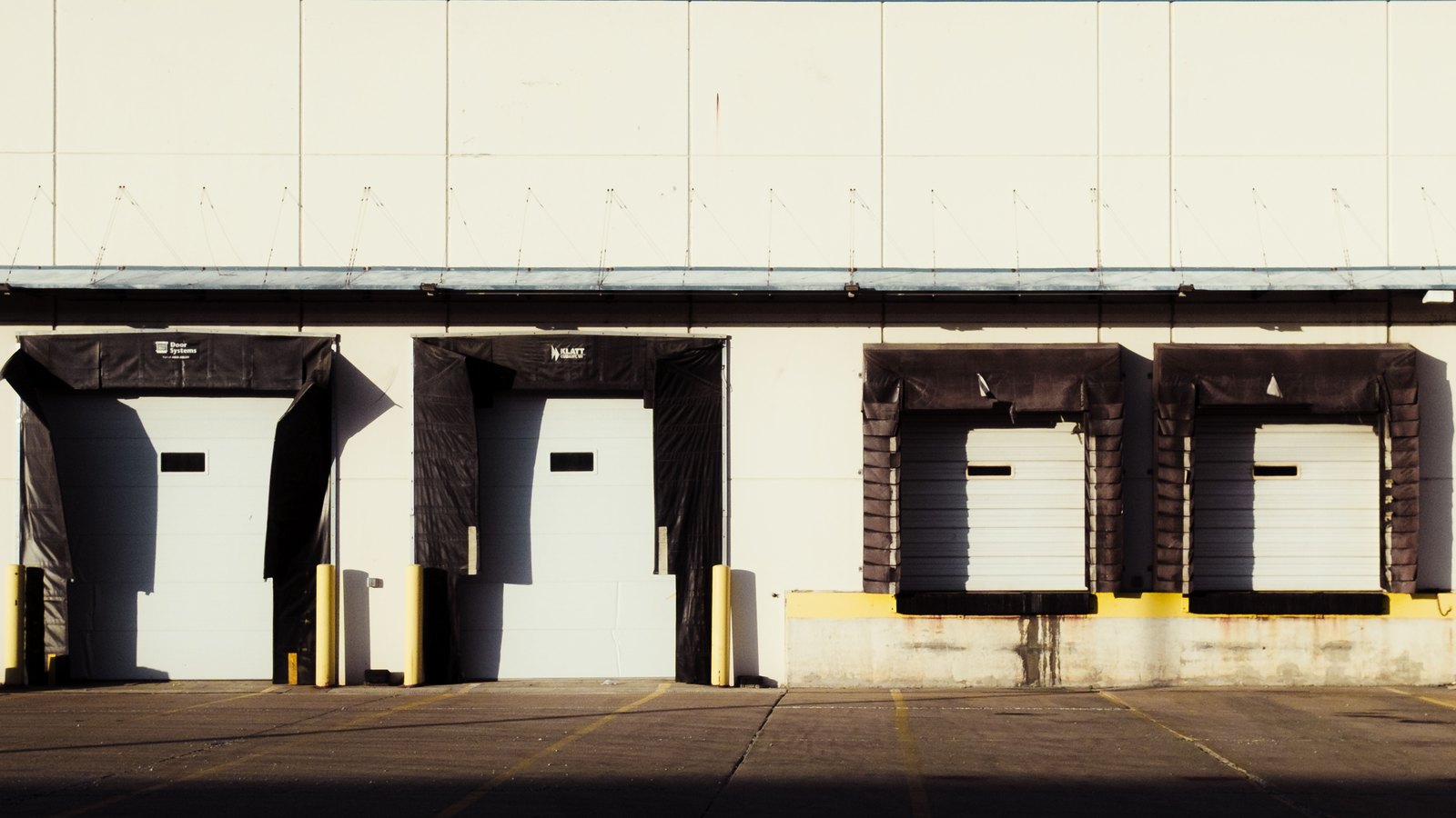 Four closed loading dock doors along a plain pale warehouse wall photographed straight on, two of them framed by dark fabric dock seals, with an empty concrete apron across the foreground in low sunlight.