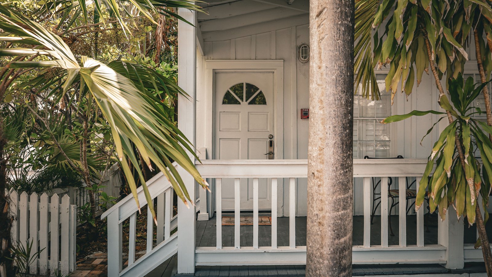 The front porch of a white clapboard house, a panelled door with a fanlight behind a white picket railing, palms in the yard.
