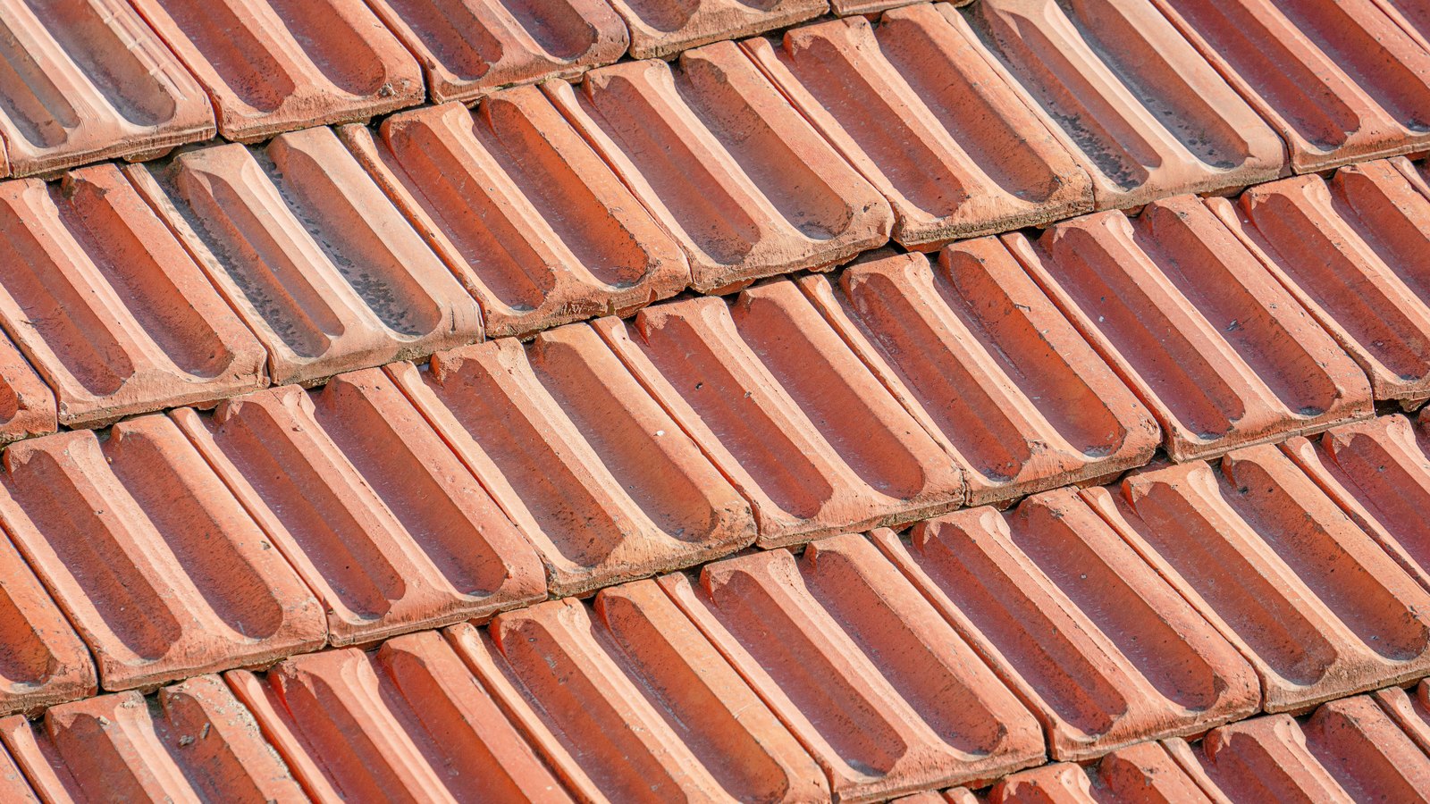 A close photograph of a tiled roof filling the whole frame, rows of ribbed terracotta pantiles overlapping in staggered courses and weathered to varying shades of red and buff. No text, brand mark, person, sky or building outline is in view.