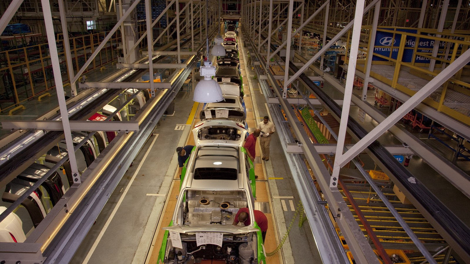 The final assembly line at Hyundai Motor Manufacturing Alabama outside Montgomery, seen from a gantry looking down the line. Car bodies in silver and white sit on green carriers with their bonnets open and build sheets taped to the front, and three workers are at the nearest two vehicles. A Hyundai banner hangs from the mezzanine on the right. Photographed for the Library of Congress in March 2010.