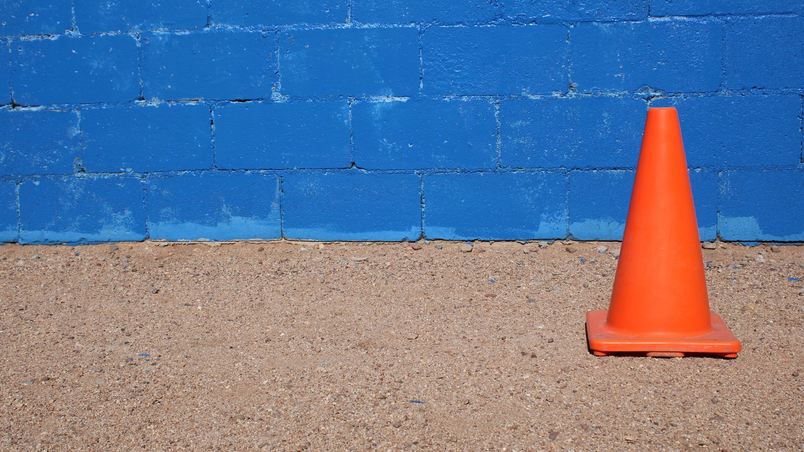 A single orange traffic cone stands on coarse sandy ground against a wall of bright blue painted concrete blocks, filling the right of the frame with empty wall and ground to the left. No people, vehicles, number plates, signs, agency markings, text or place names appear anywhere in the frame. Stock photograph, not a photograph of this event.